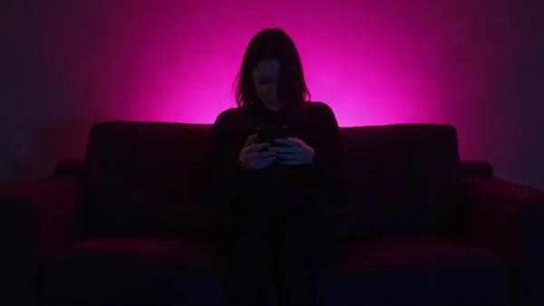 Silhouet op de rug gezien op een bank met een telefoon, in een donkere kamer met magenta gloed (illustratie bij het artikel)